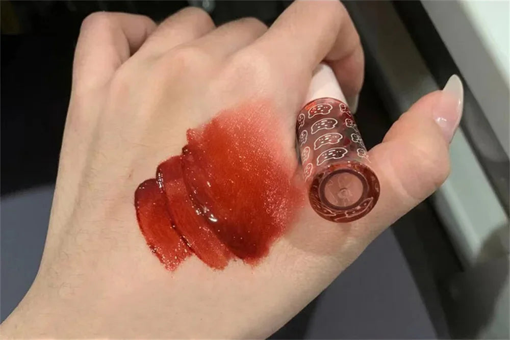 Hand with glossy red lip tint swatch next to a cute patterned tube, beauty makeup product