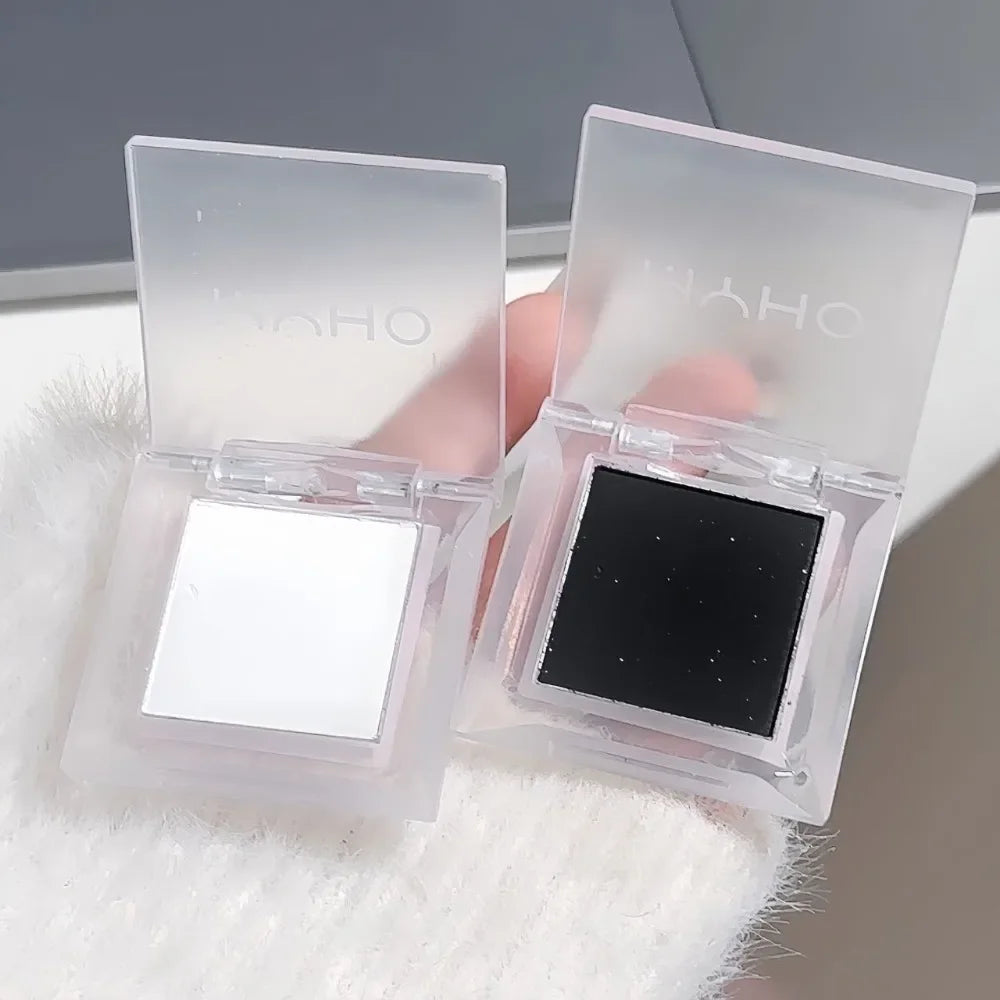 Hand holding two open eyeshadow compacts, one with white and one with black eyeshadow.